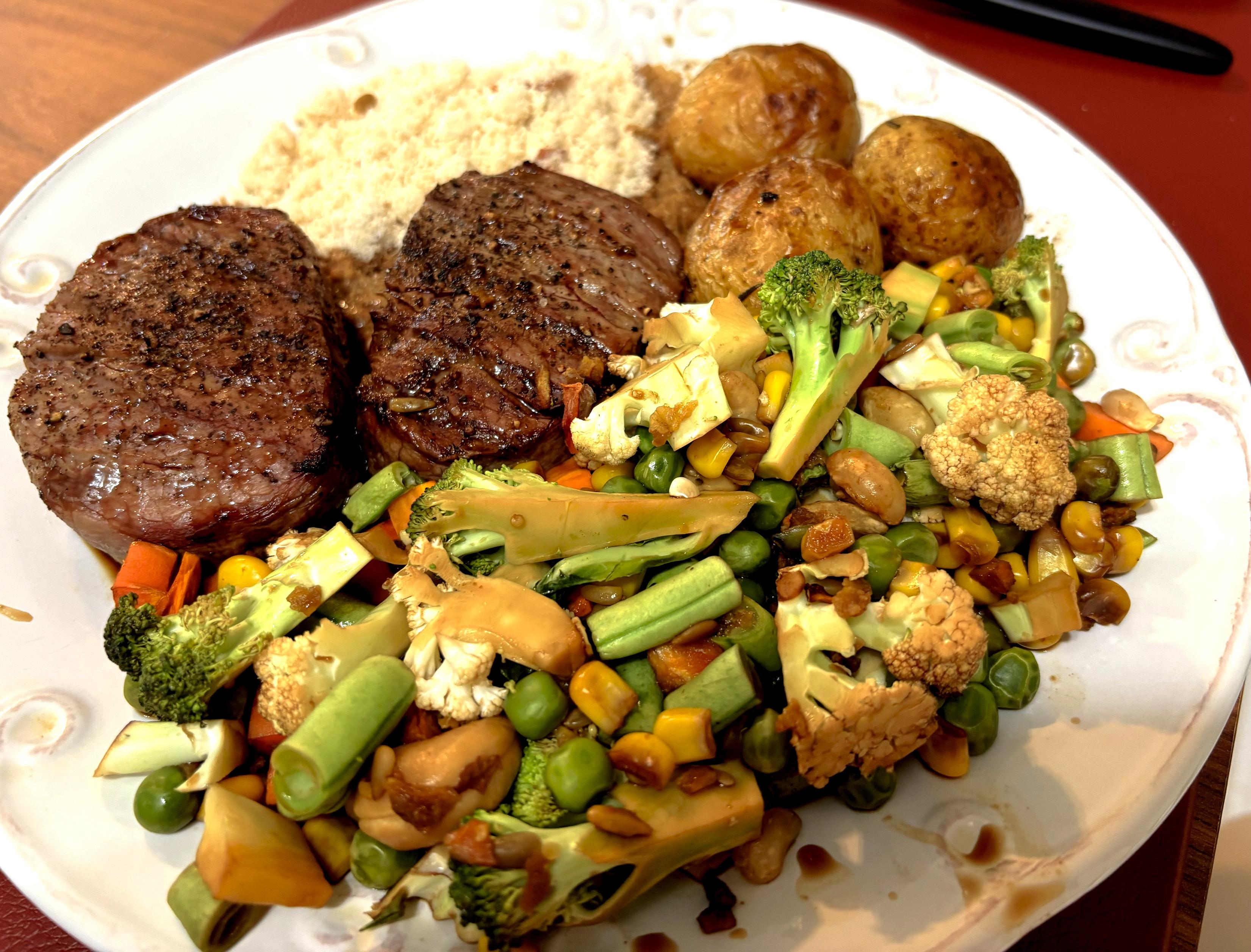 Um prato servido contendo farofa, 3 batatas portuguesas coradas, grande quantidade de legumes picados, e 2 bifes cortados tipo medalhão. 