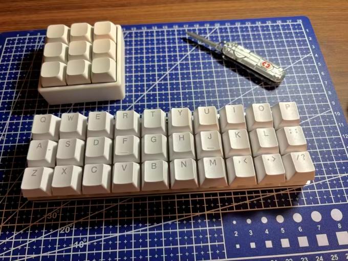 Um teclado de 30 teclas, apenas com as teclas de letras, vírgula, ponto, / e ponto e vírgula, ao lado de um teclado de 3x3 teclas sem rótulos, e de um canivete que calhou de estar na bancada na hora da foto.