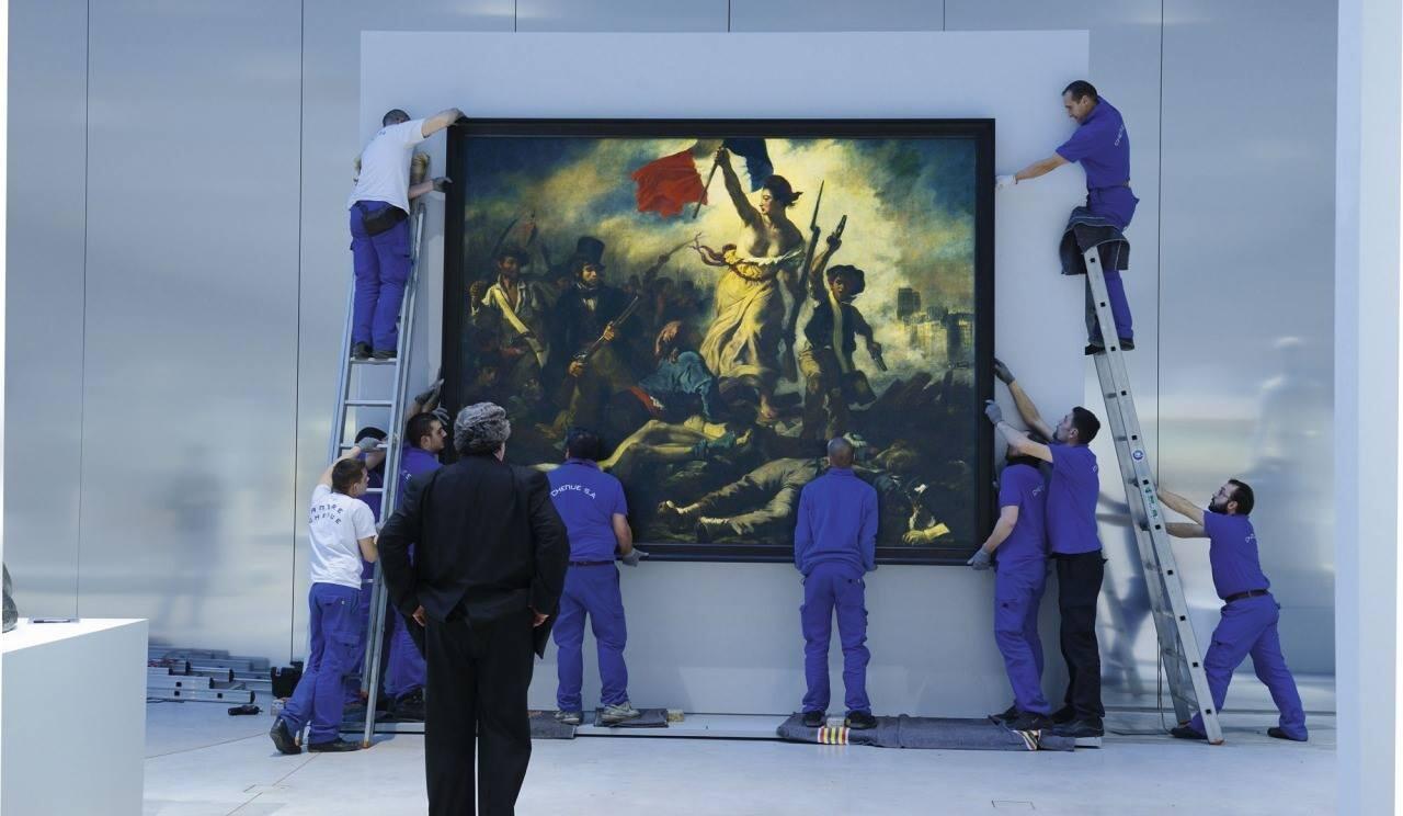 Foto de uma pessoa de paletó escuro que observa, com as mãos nos bolsos, enquanto 10 pessoas uniformizadas penduram um quadro símbolo da revolução francesa.