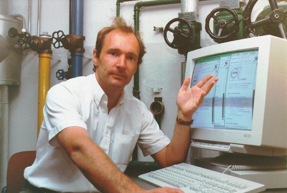 Foto do jovem Tim Berners-Lee, de camisa social de manga curta, num ambiente cheio de canos e válvulas aparentes na parede, junto a uma workstation rodando o seu software para acessar o site do próprio CERN.