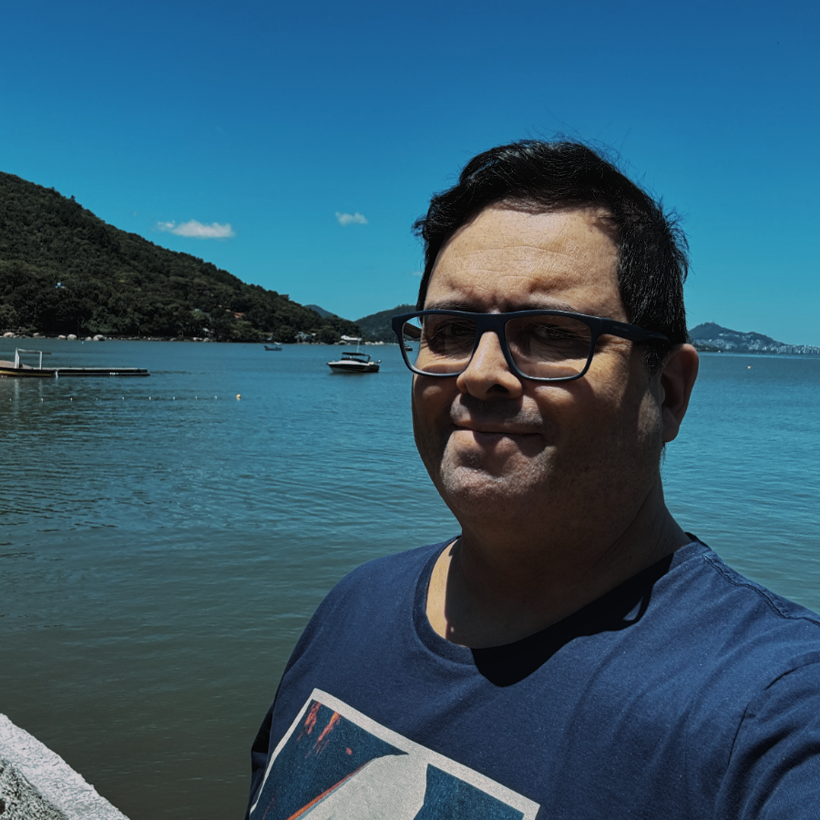 Foto minha, olhando para a câmera, de óculos e camiseta azul, tendo ao fundo o céu azul, o mar azul, e uma lancha fundeada em frente a um trapiche distante.