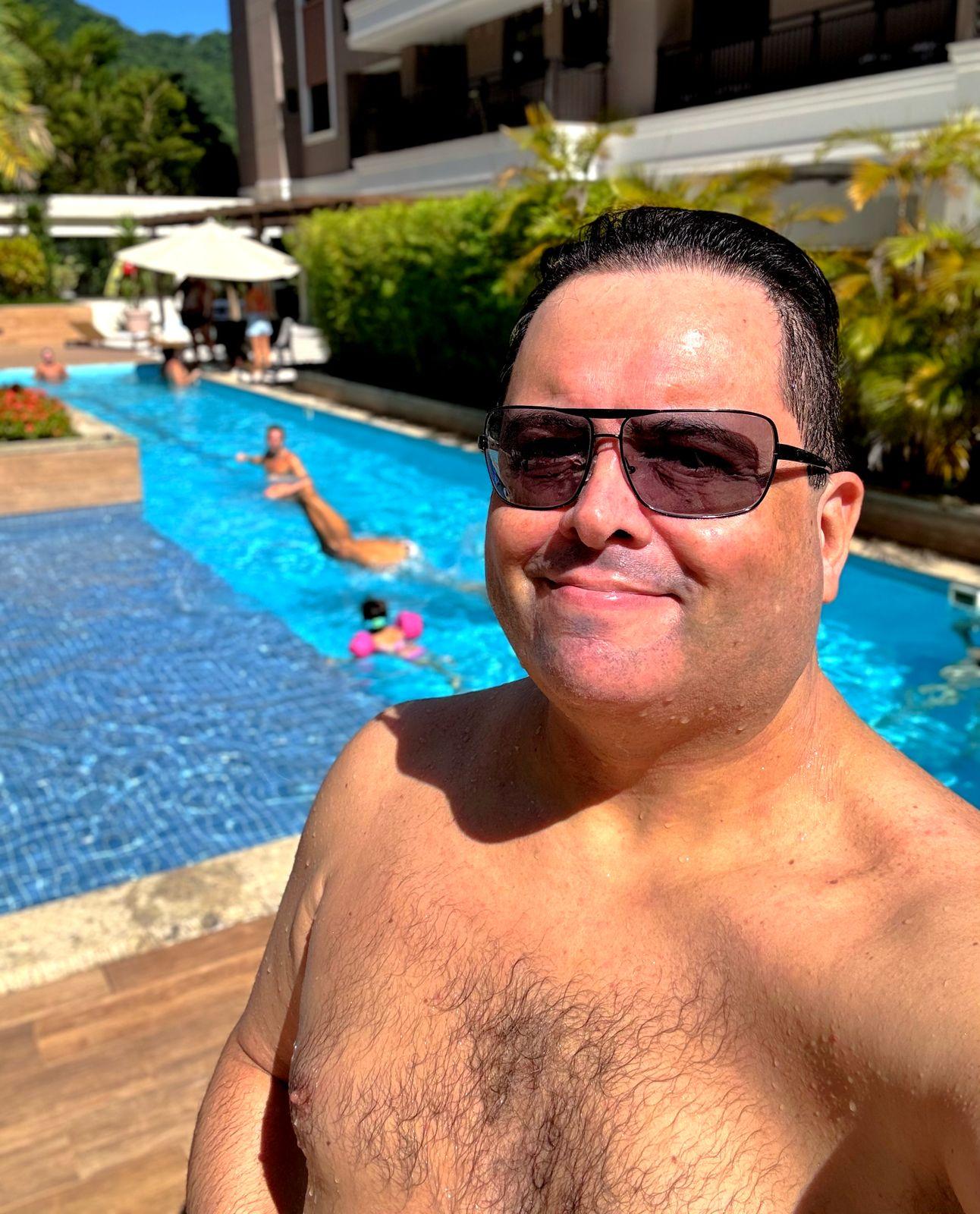 foto minha, de óculos escuros, cabelo molhado e sem camisa, ao sol, tendo uma piscina e vegetação ao fundo.