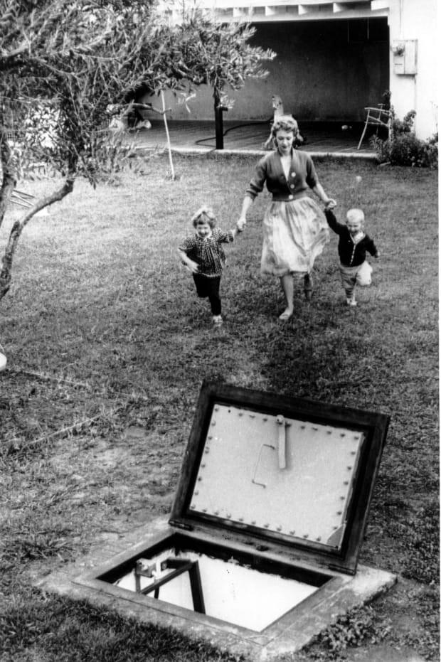 Foto em preto e branco de uma mulher correndo, levando duas crianças pelas mãos, em direção à porta aberta de um abrigo subterrâneo.