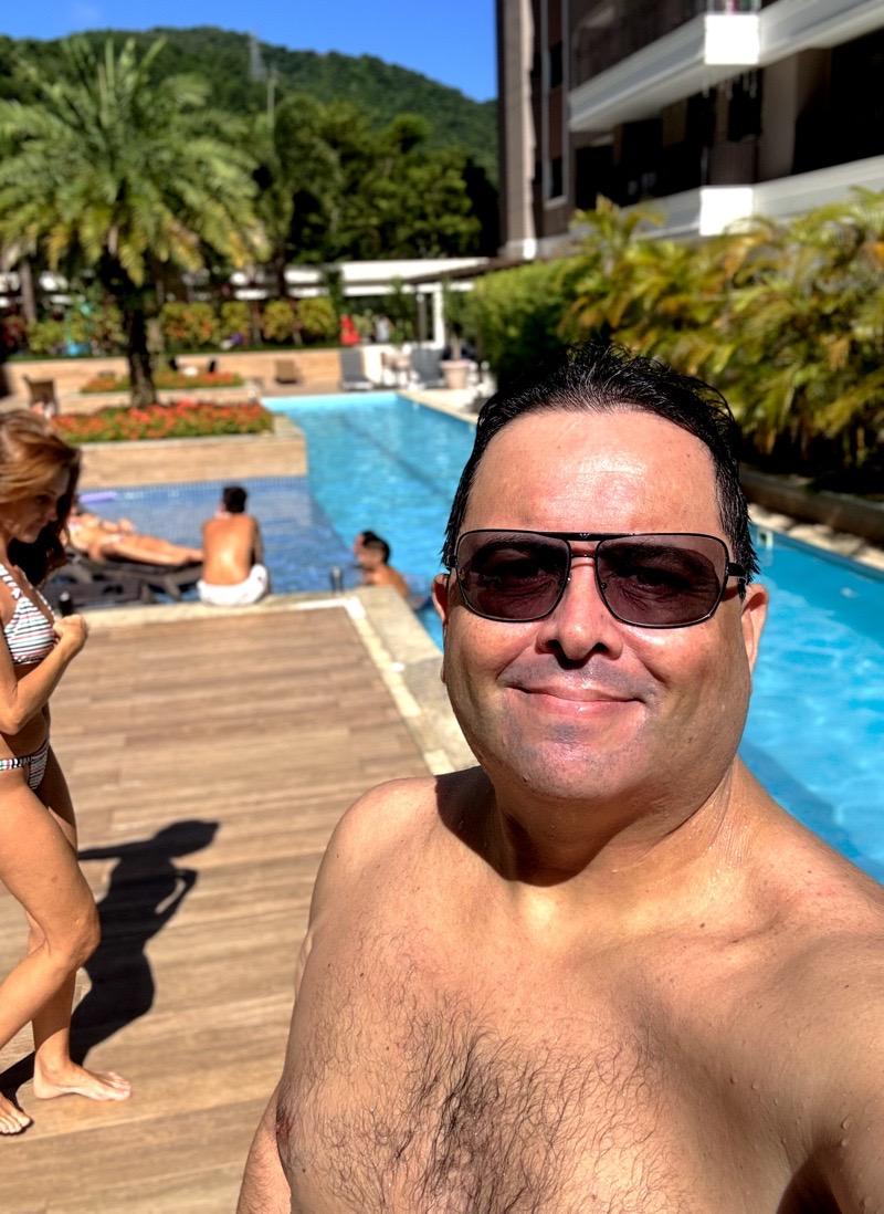 Foto minha, molhado, sem camisa e de óculos escuros, olhando para a câmera, com a piscina, vegetação e algumas pessoas ao fundo, numa cena ensolarada