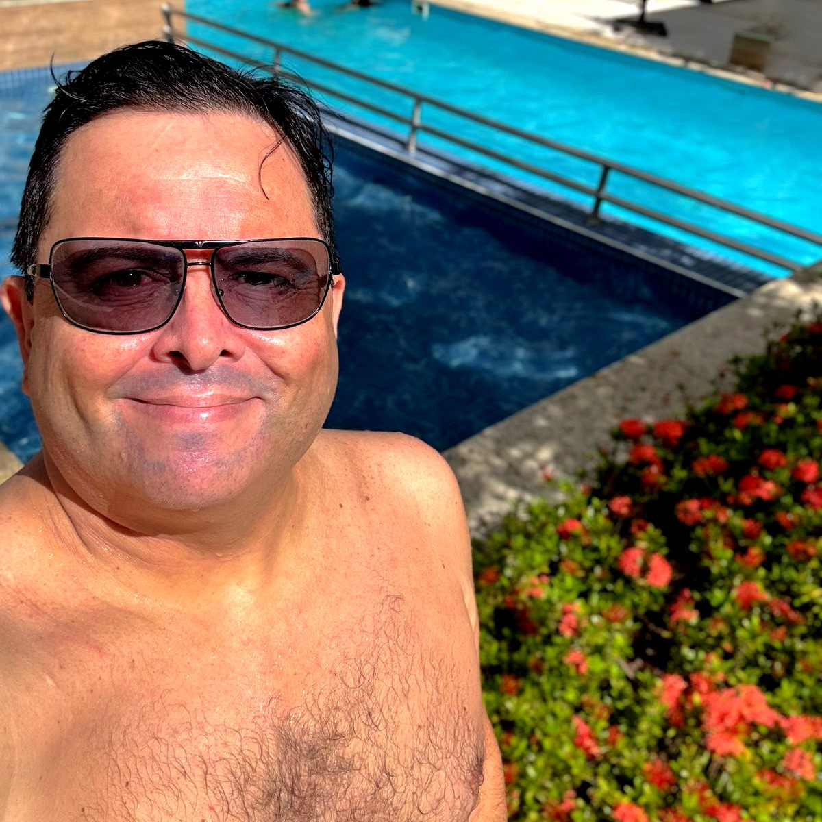foto minha, sem camisa e com os cabelos molhados, usando óculos escuros, em cena ensolarada, tendo ao fundo uma piscina e um canteiro de flores vermelhas e relva verde.