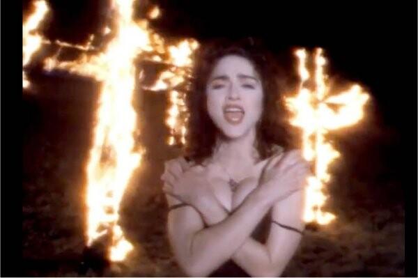 Cena do clip, com a cantora Madonna, braços cruzados sobre o peito, à frente de várias cruzes em chamas