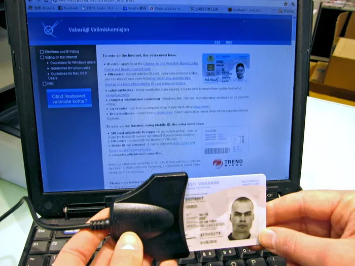 Foto de um documento de identidade da Estônia, conectado a um leitor, em frente a uma tela de notebook mostrando a versão em inglês do site de votação on-line da Estônia na década retrasada.