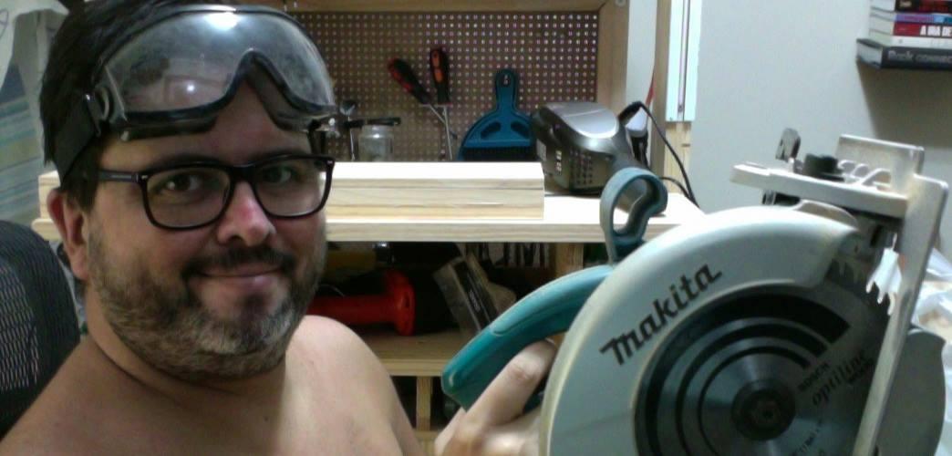 foto minha, com barba e sem camisa, com o óculos de proteção temporariamente descansando sobre a testa, sorrindo para a câmera e segurando uma serra circular Makita, tendo ao fundo minha bancada de marcenaria.