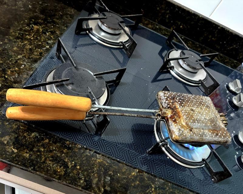 Uma sanduicheira tipo Tostex, de alumínio, com cabo de madeira e evidentes marcas de uso ao longo dos anos. Ela está em uma boca de fogão, acesa.