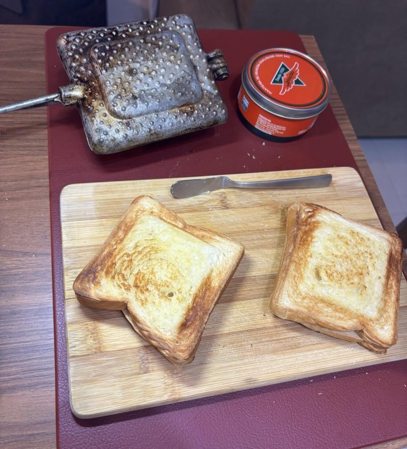 Dispostos em uma tábua, 2 sanduíches tostados. Ao redor da tábua, uma lata de manteiga, e uma sanduicheira tipo Tostex.