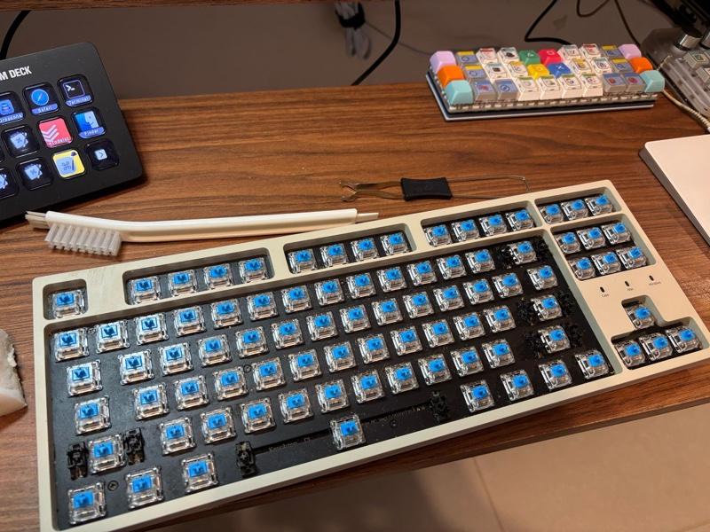 Foto do meu teclado na escrivaninha, sem as teclas, com a limpeza em andamento, ao lado de uma escova e de uma ferramenta para facilitar a extração de teclas