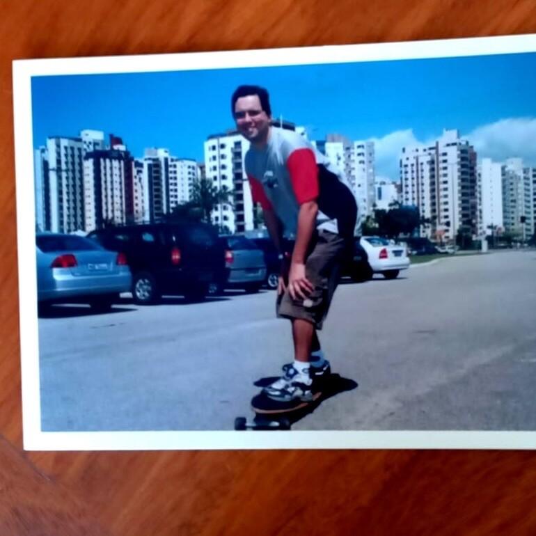 Foto antiga presa em um mural. Estou no asfalto, sobre um skate longboard, de camiseta colorida e bermuda, sorrindo e olhando para a câmera, com os prédios e os carros estacionados ao fundo.