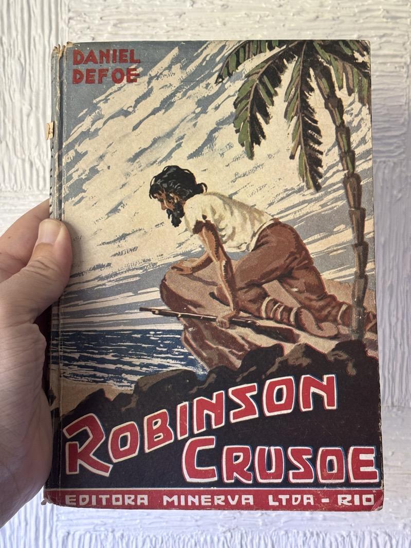 Capa com ilustração colorida do personagem náufrago, de barba e cabelo compridos, usando uma pedra para se ocultar parcialmente enquanto observa um cenário praiano, entre pedras e ao lado de uma palmeira