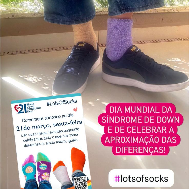 Foto dos meus pés, usando tênis azul, com a barra da calça erguida para mostrar que uma meia é roxa e a outra é marrom. A foto inclui um banner da campanha Lots Of Socks, e o texto: Dia Mundial da Síndrome de Down e de Celebrar a Aproximação das Diferenças