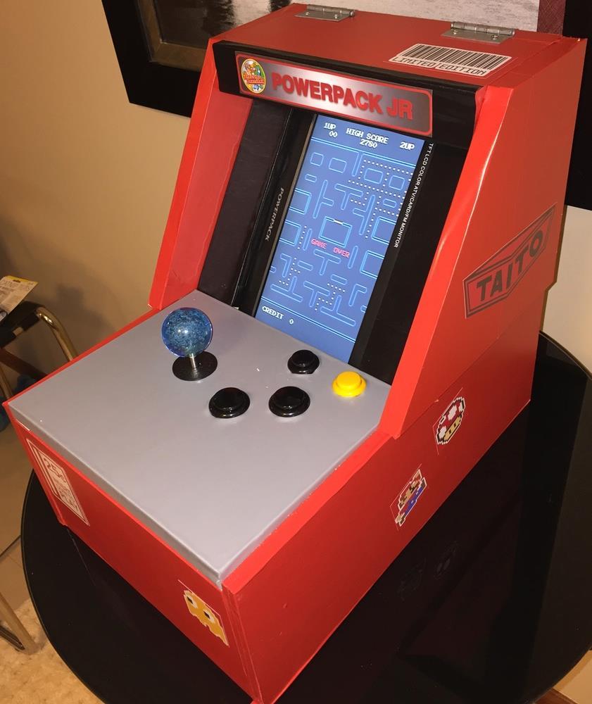 Uma mini-máquina de fliperama, de mesa, com cerca de 60cm de altura, alavanca de comando, 4 botões, tela vertical, com acabamento em cores vermelho, reto e cinza, e adesivos alusivos à Taito e a personagens de jogos dos anos 80. A tela dela nostra o jogo Pac-Man, da Namco.