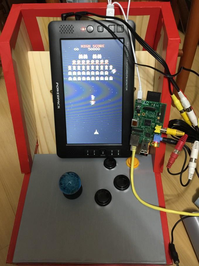 Uma mini-máquina de fliperama, de mesa, com cerca de 60cm de altura, alavanca de comando, 4 botões, tela vertical, com o gabinete parcialmente aberto, permitindo ver a fiação, o mini-monitor de vídeo em seu interior, e a placa Raspberry Pi que a controla.