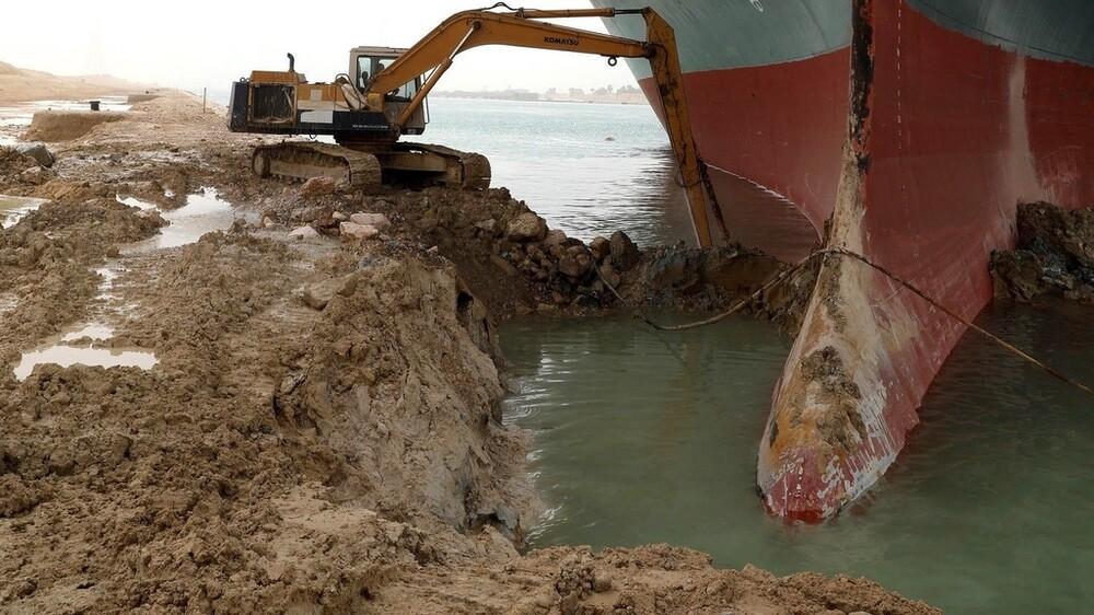 Proa do navio Ever Given junto à margem do canal, e uma escavadeira operando ao seu lado.