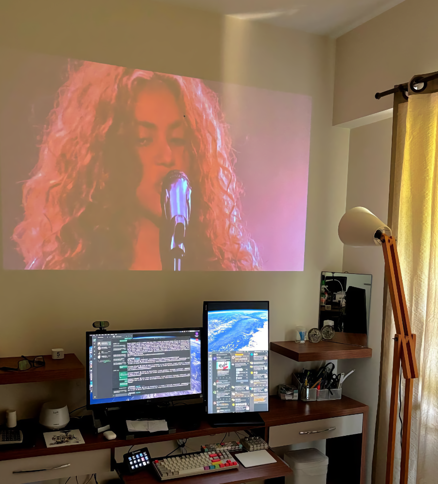 Foto do meu escritório, com uma projeção acima dos 2 monitores. A projeção, feita diretamente na parede, mostra o rosto da cantora Shakira, em close, em frente a um microfone de pedestal.