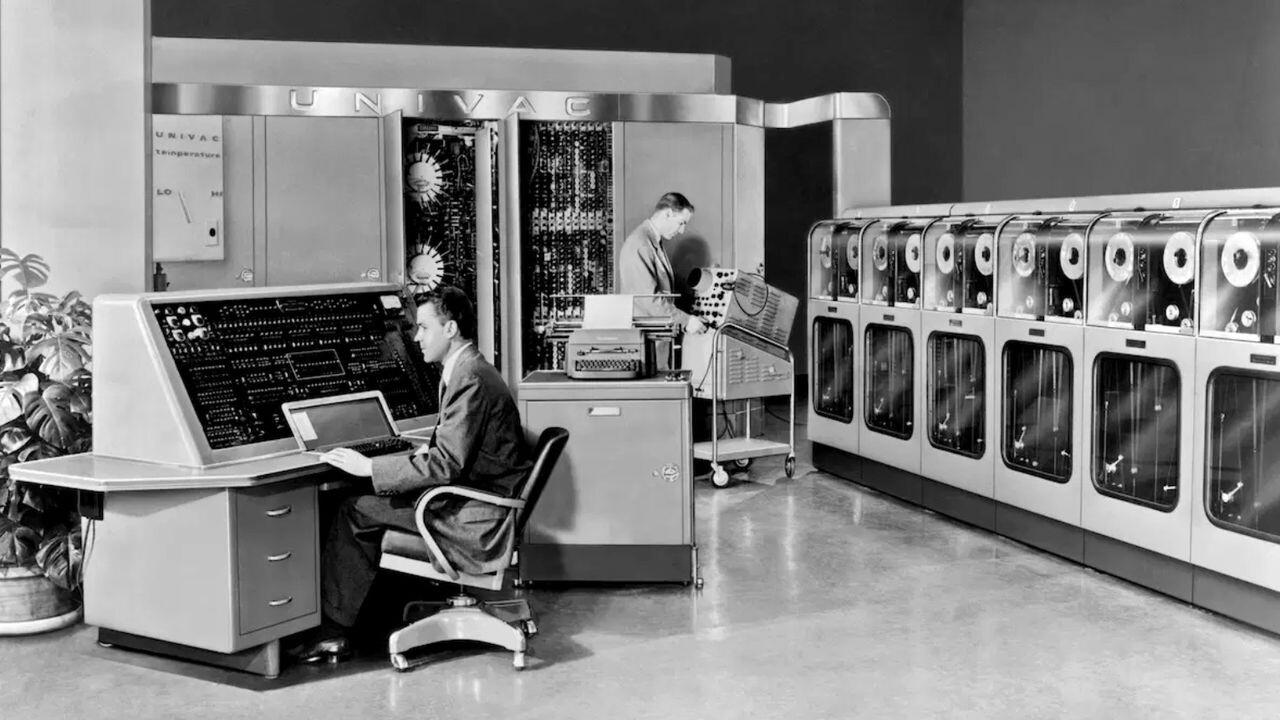 Foto em preto e branco de parte do Univac, com 2 operadores, um deles sentado junto ao console.